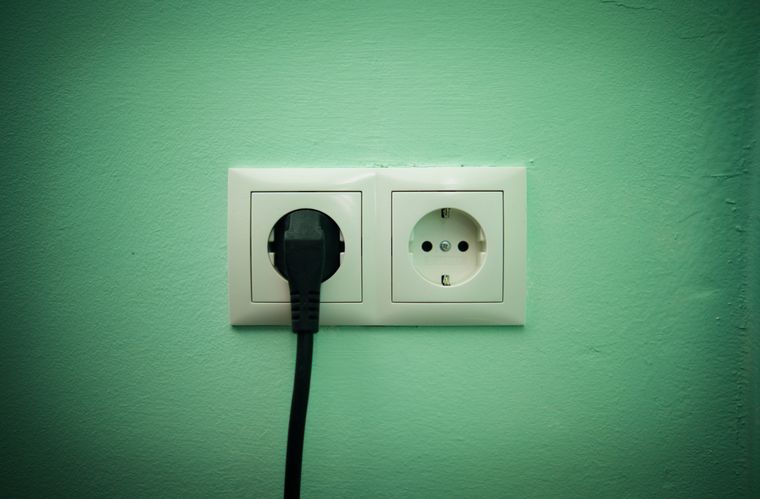 Eine Doppelsteckdose vor einer grünen Wand. Der rechte Stecker ist frei, die linke Dose ist von einem schwarzen 
Stecker belegt.