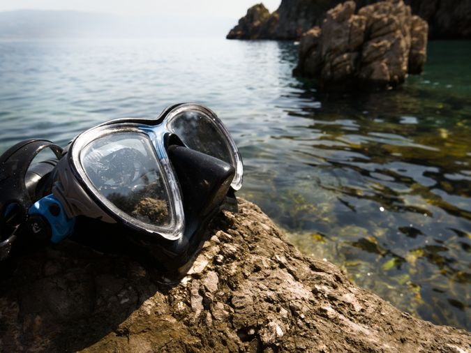 Eine Tauchermaske liegt auf einem Stein der Küste, dahinter sieht man unscharf das Meer