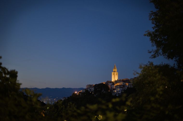 Die Skyline einer kroatischen Kleinstadt zur blauen Stunde, die Szene ist umrandet von Baumwerk. Der Kirchturm 
sticht mit seiner gelben Beleuchtung aus der dunklen, blauen Szene hervor