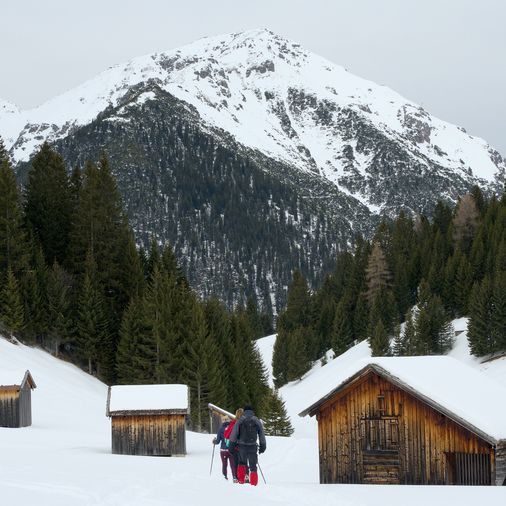 3 Schneeschuhwanderer, die zwischen 2 Holzhütten durchlaufen. Im Hintergrund ist ein Berggipfel zu sehen, auf den
sie zulaufen
